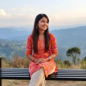 Young woman in vibrant kurti sitting on a bench with a scenic mountain backdrop, smiling joyfully.