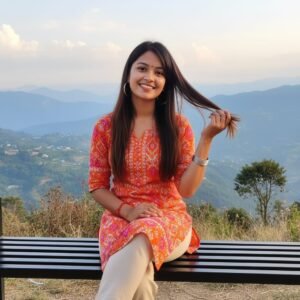Young woman in vibrant kurti sitting on a bench with a scenic mountain backdrop, smiling joyfully.